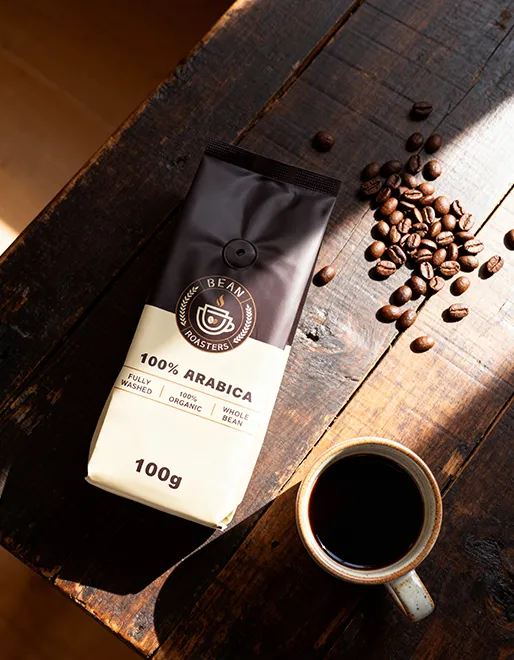 A bag of Bean Roasters 100% Arabica coffee on a dark, rustic wooden table next to a cup of black coffee and scattered whole beans.